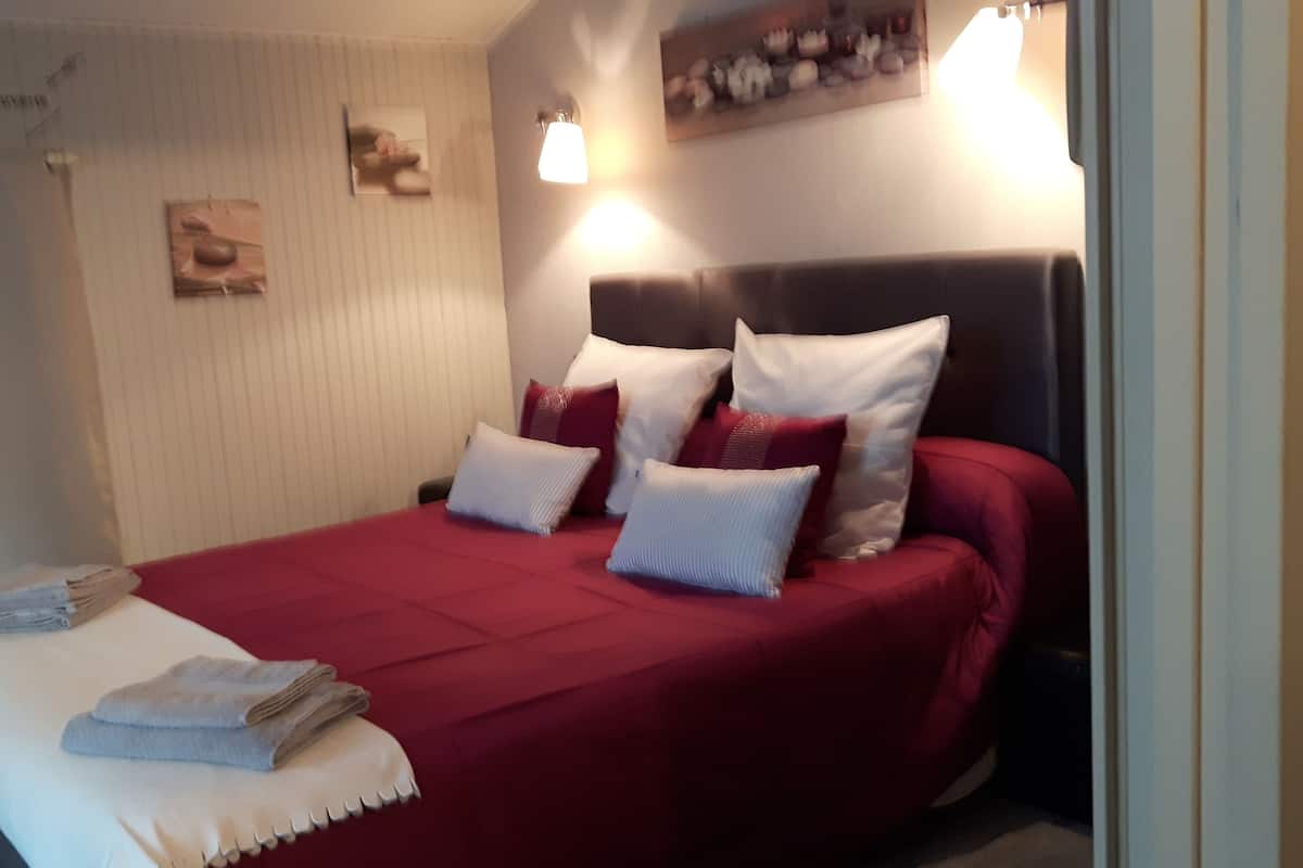 A neatly made bed with a red bedspread and white pillows, a small bedside table with a folded towel, and a wall-mounted light fixture.