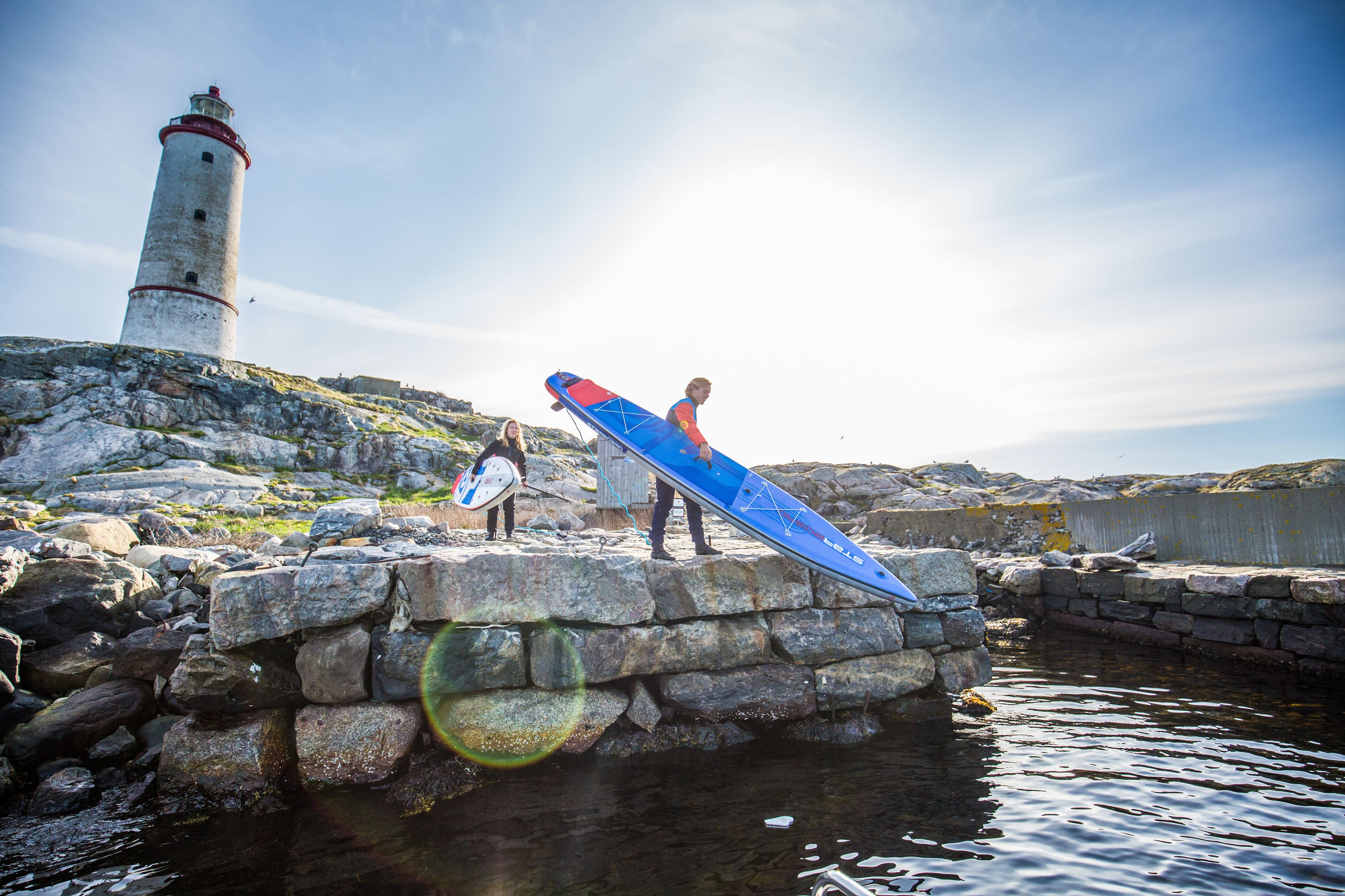 A lighthouse, a rocky shoreline, and a person carrying a blue surfboard.