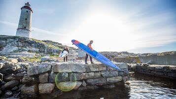 A lighthouse, a rocky shoreline, and a person carrying a blue surfboard.