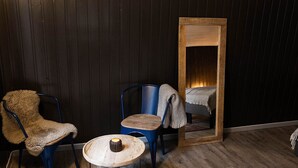 A room with a wooden floor, a fur chair, a blue chair, a small wooden table with a candle, and a mirror reflecting a bed with a blanket. - Raet Camping (Arendal)
