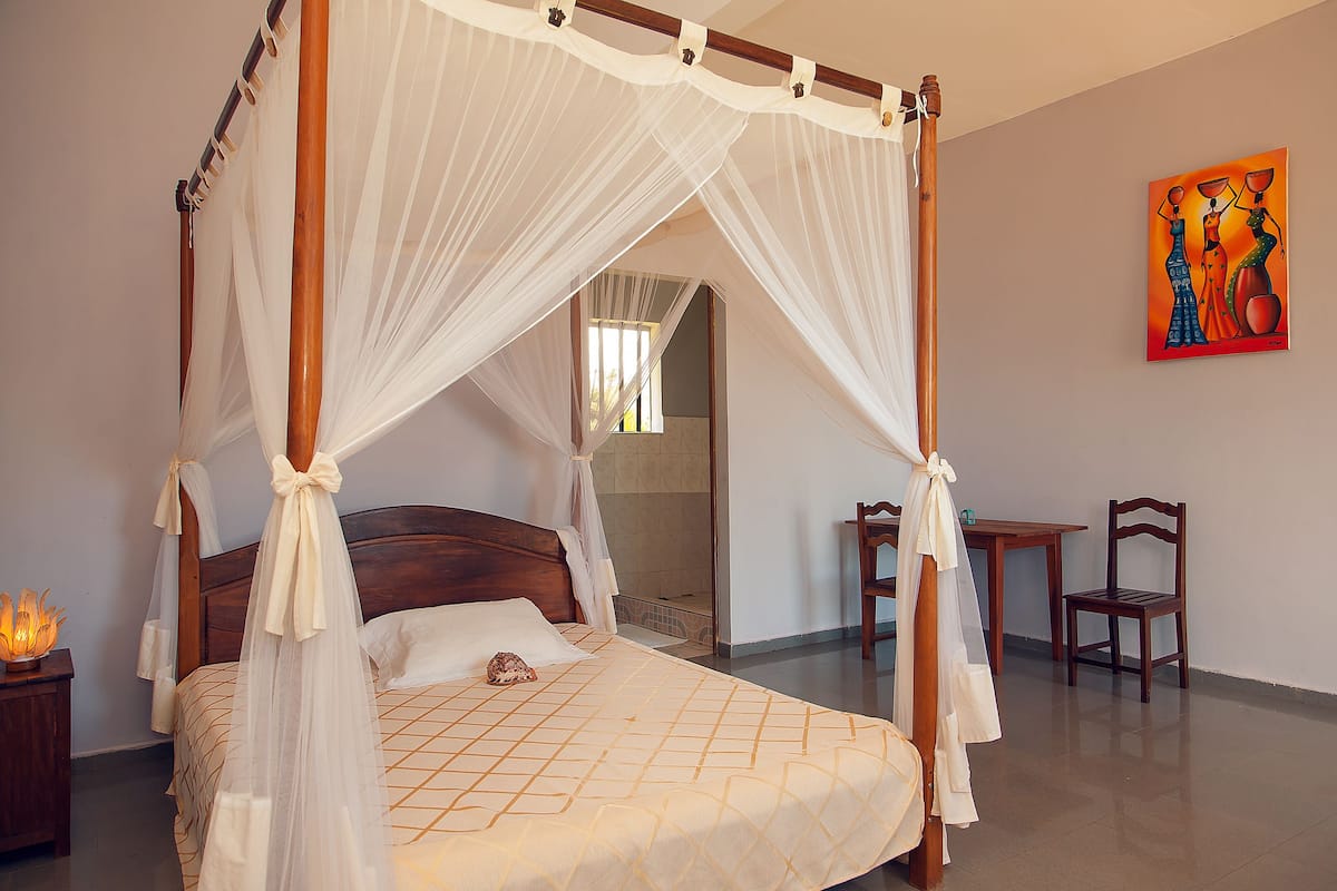A four-poster bed with a canopy, a wooden nightstand with a lamp, a wooden chair, and a table with a chair.