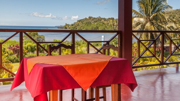 Une table recouverte d’une nappe rouge, placée sur un balcon d’où l’on peut admirer un paysage magnifique sur l’océan et une végétation luxuriante.