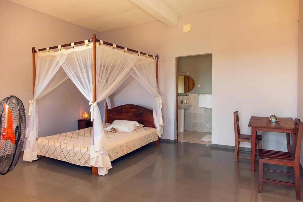 A four-poster bed with a canopy, a bedside table with a lamp, a wooden chair, and a small table with a decorative item.
