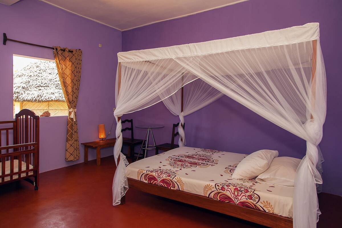 A room with a bed under a white canopy, a wooden chair, a small table with a lamp, and a window with a curtain.