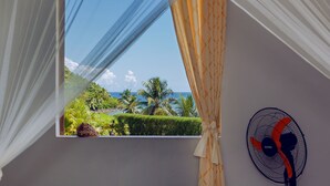 Une chambre avec une fenêtre donnant sur un paysage tropical, un ventilateur et un rideau.