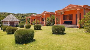Un complexe hôtelier composé de plusieurs bâtiments aux façades orange, de haies soigneusement taillées et d’une structure surmontée d’un toit de chaume.