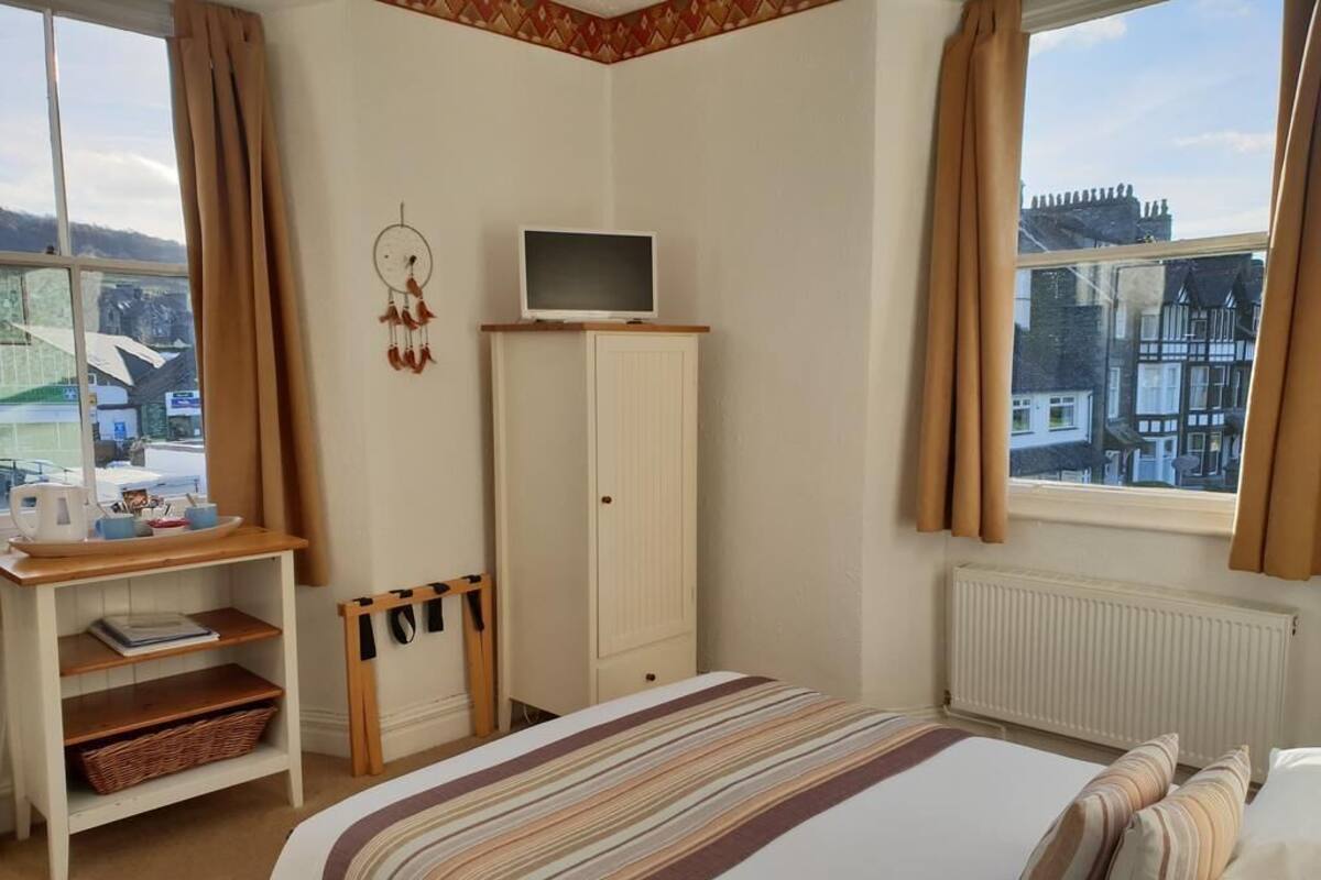 Ein Schlafzimmer mit einem Bett, einem Schreibtisch, einem Fernseher auf einer Kommode, einem Fenster mit Vorhängen und Blick auf Gebäude.