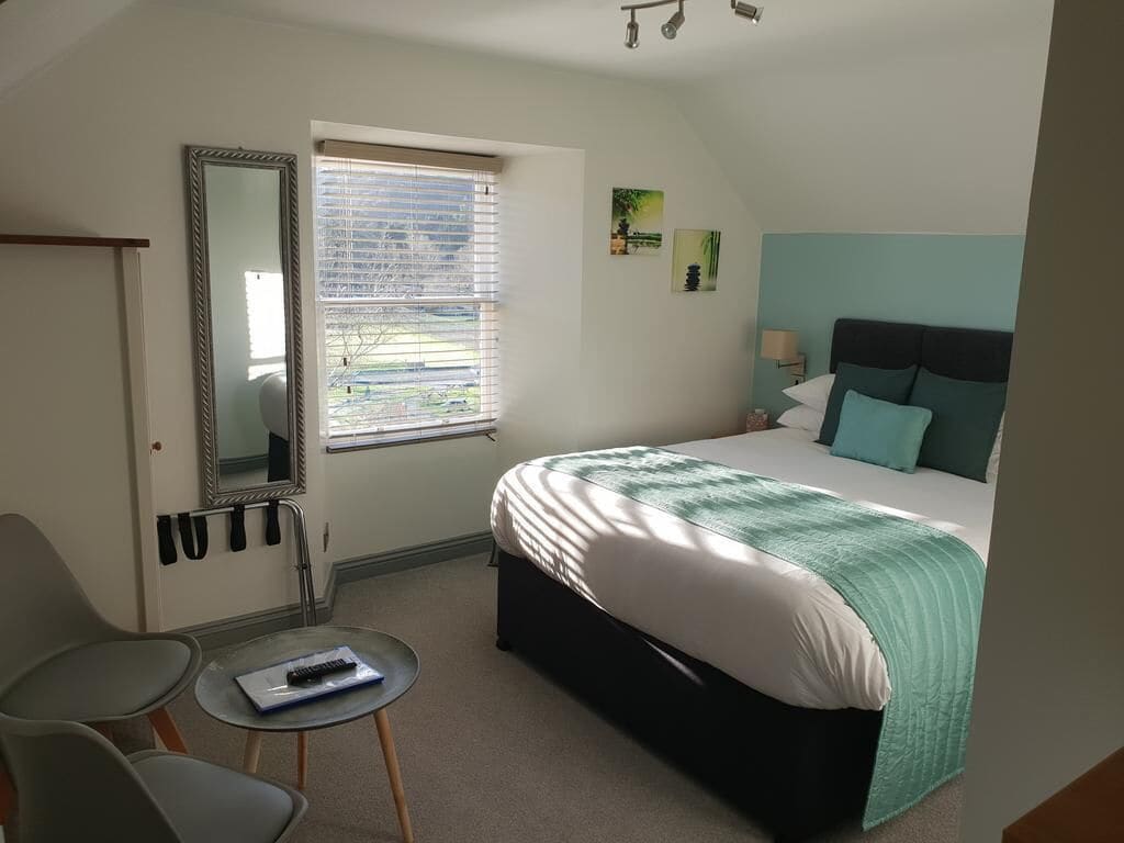 A bedroom with a bed, a chair, a small table, a window with blinds, a mirror, and a wall-mounted lamp.