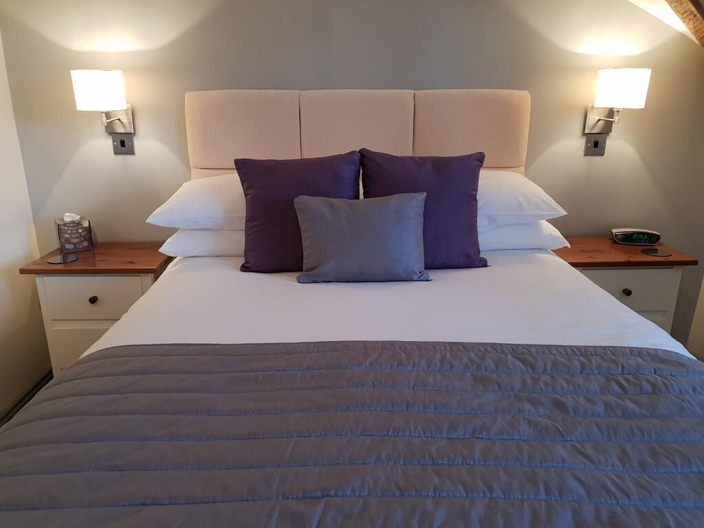 A neatly made bed with a grey duvet, two white pillows, and three purple pillows. There are two bedside tables with lamps and a clock.