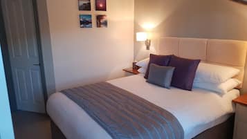 Una cama bien hecha con una manta gris, una lámpara de noche y una mesita de noche con lámpara. Hay tres cuadros enmarcados en la pared.