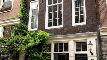 Een klassiek bakstenen gebouw met meerdere ramen en een zwarte deur, omgeven door potplanten en een bankje.