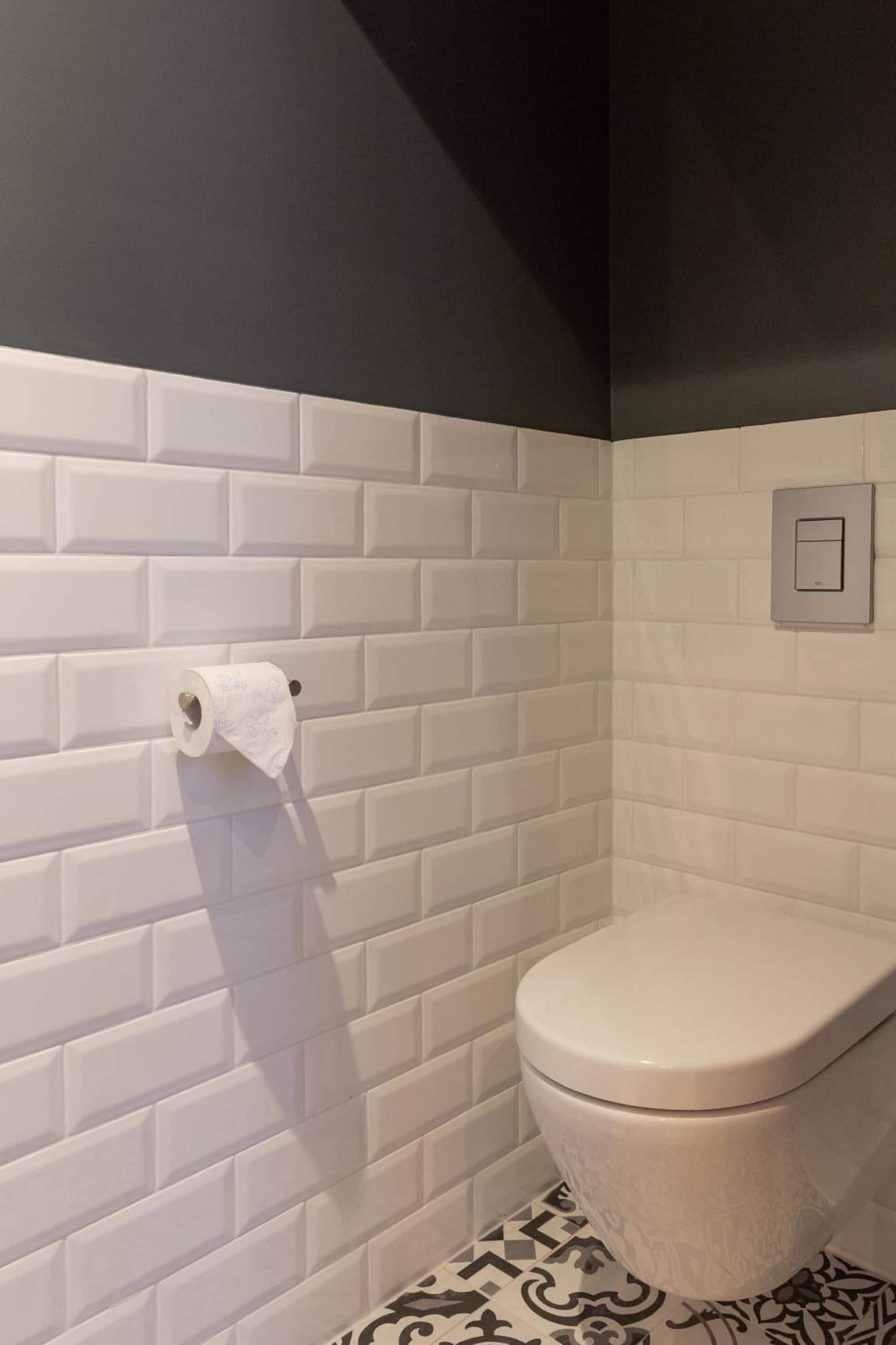 Casa de banho com paredes em azulejos brancos tipo metrô, um vaso sanitário e um rolo de papel higiénico na parede.