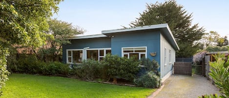 A single-story house with a blue exterior, a front yard with a well-maintained lawn, and a gravel driveway leading to the entrance.