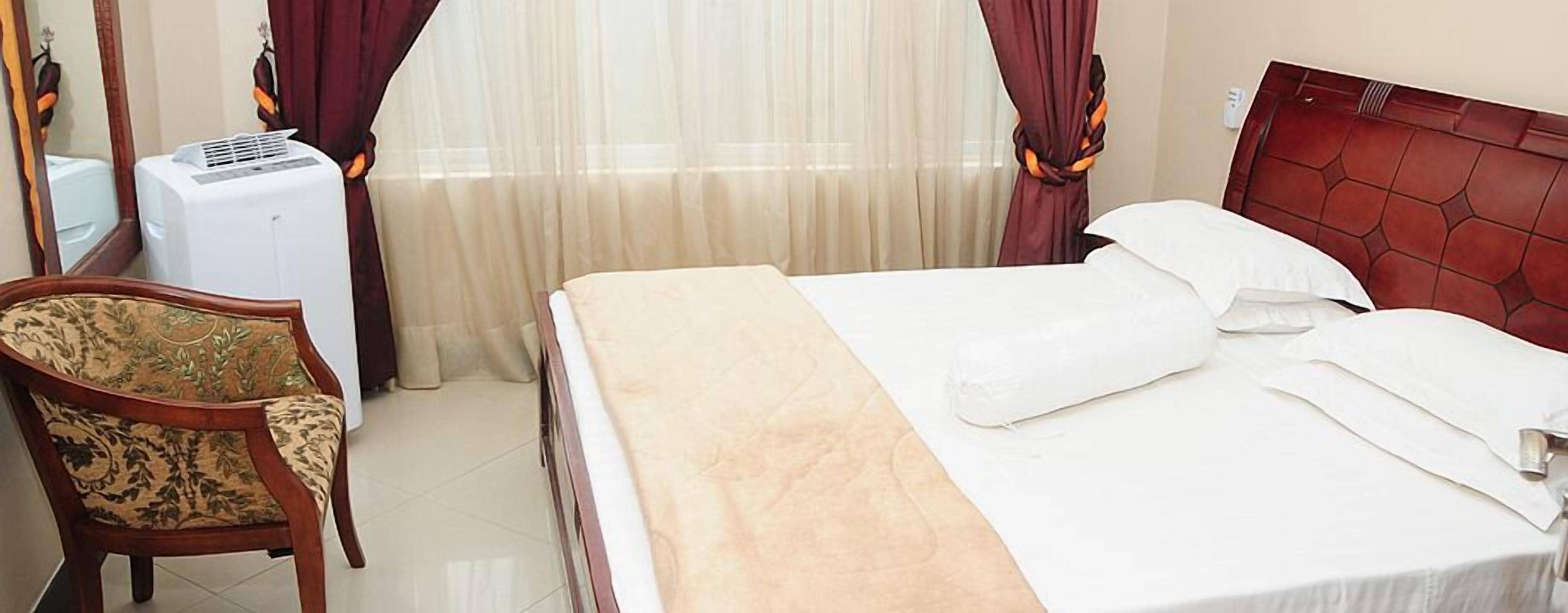 A hotel room with a bed, a chair, a window with curtains, and an air conditioning unit.
