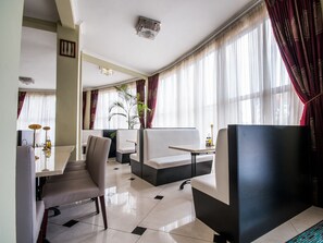 A modern hotel room with a dining area, a sofa, a TV, and a large window with curtains.