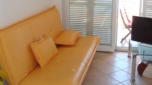 Ett rum med en gul soffa, en tv och en skjutdörr i glas som leder ut till ett utomhusområde.