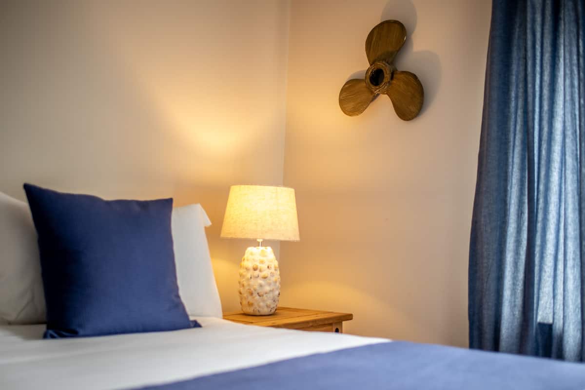 A neatly made bed with a blue pillow, a lamp on a wooden bedside table, and a wall-mounted fan.