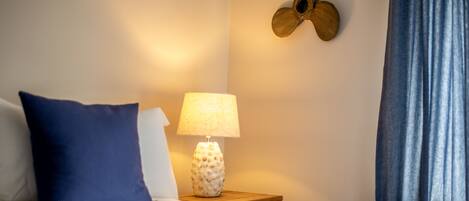 A neatly made bed with a blue pillow, a lamp on a wooden bedside table, and a wall-mounted fan.
