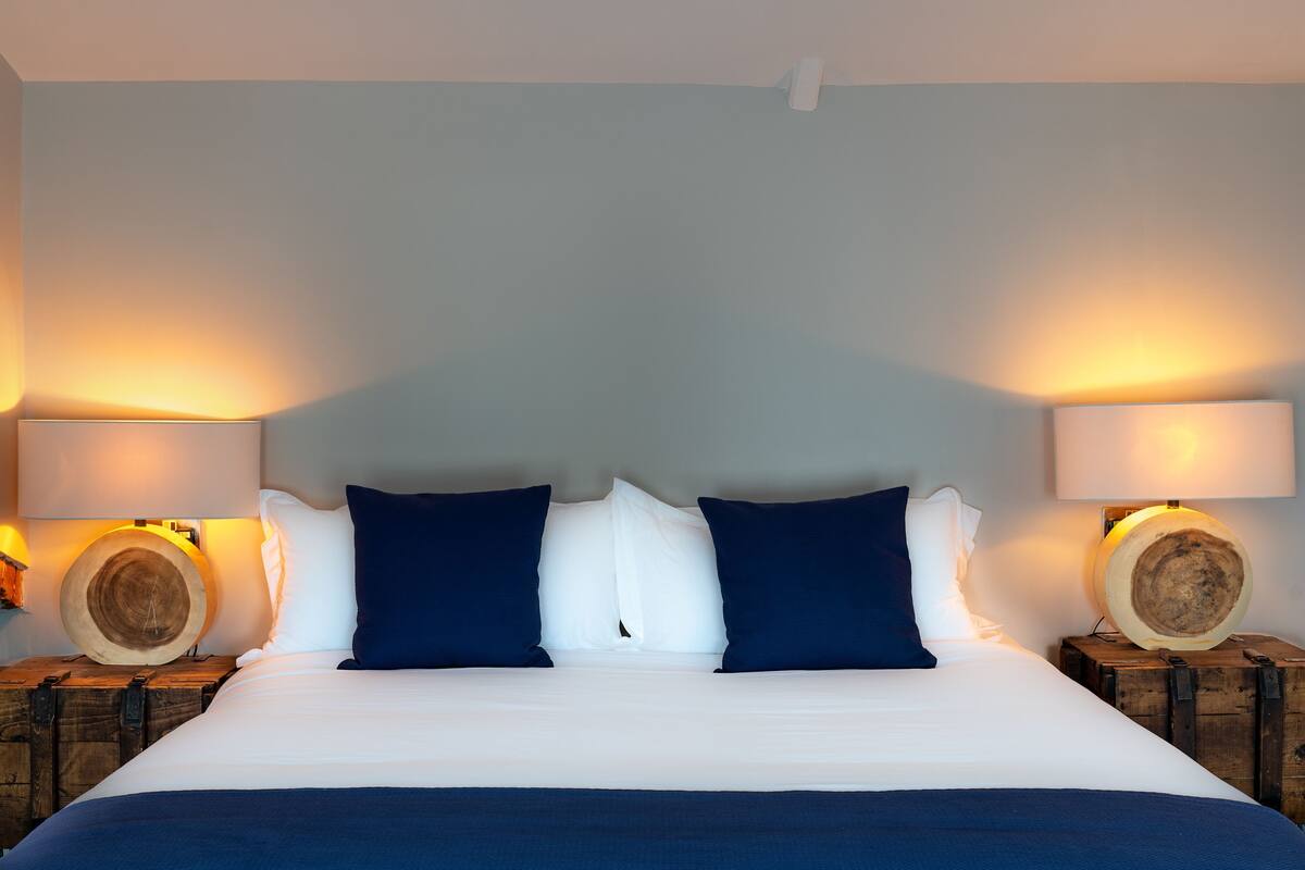 A bed with white linens, two dark blue pillows, and two round wooden nightstands with lamps.