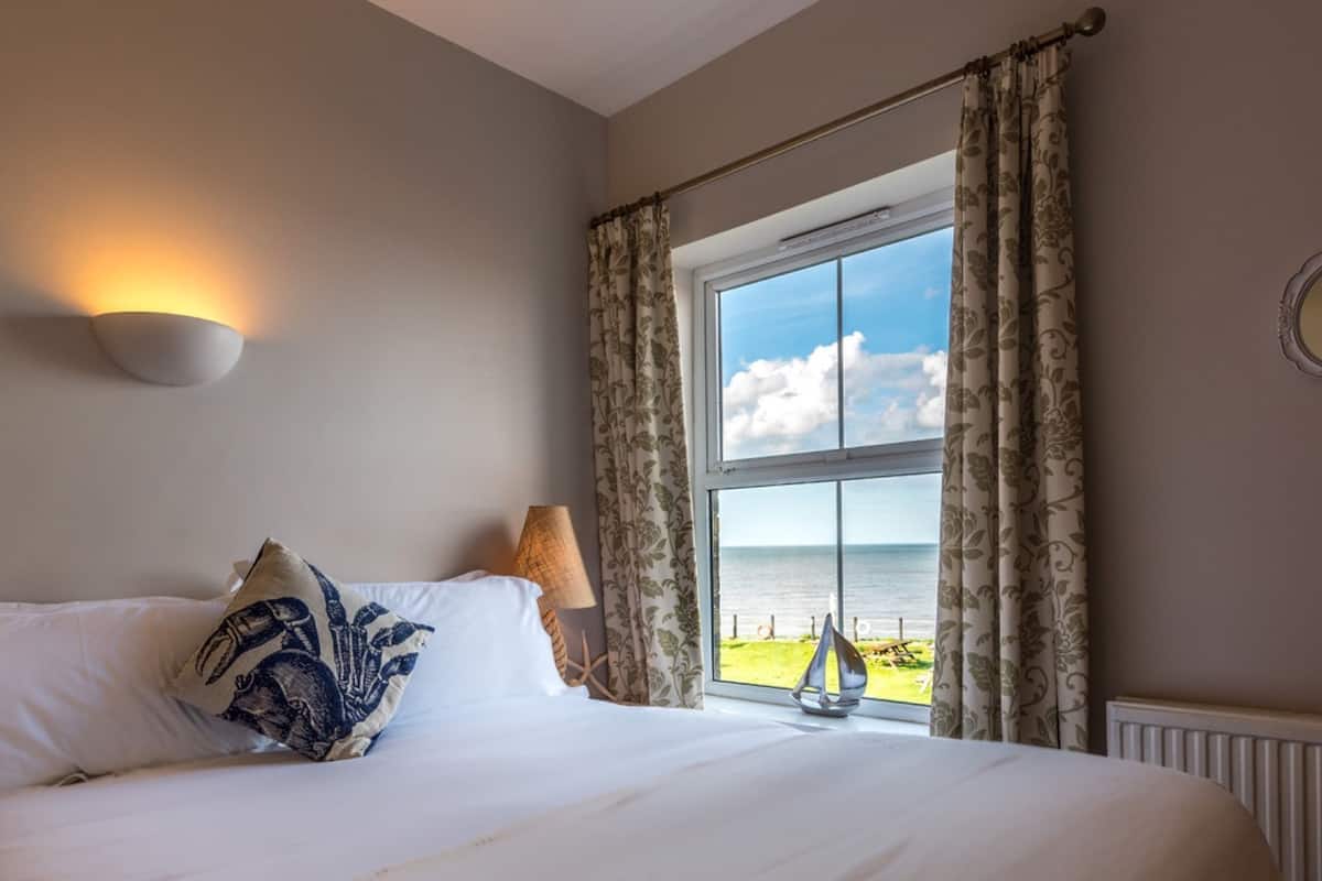 A bedroom with a bed, a window with curtains, a wall-mounted lamp, and a round mirror.