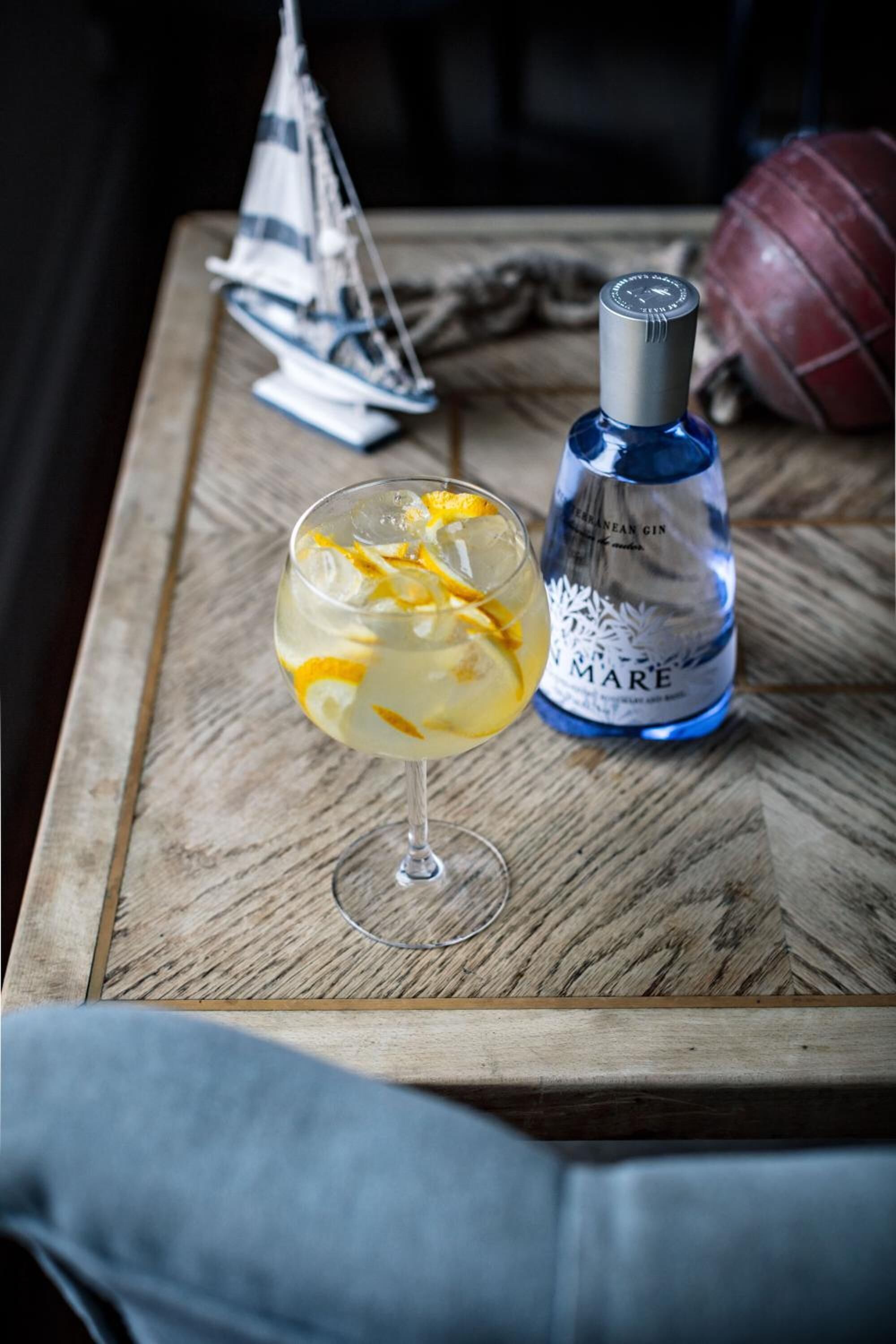 A glass of cocktail with orange slices on a wooden table, a bottle of gin, and a small sailboat model in the background.