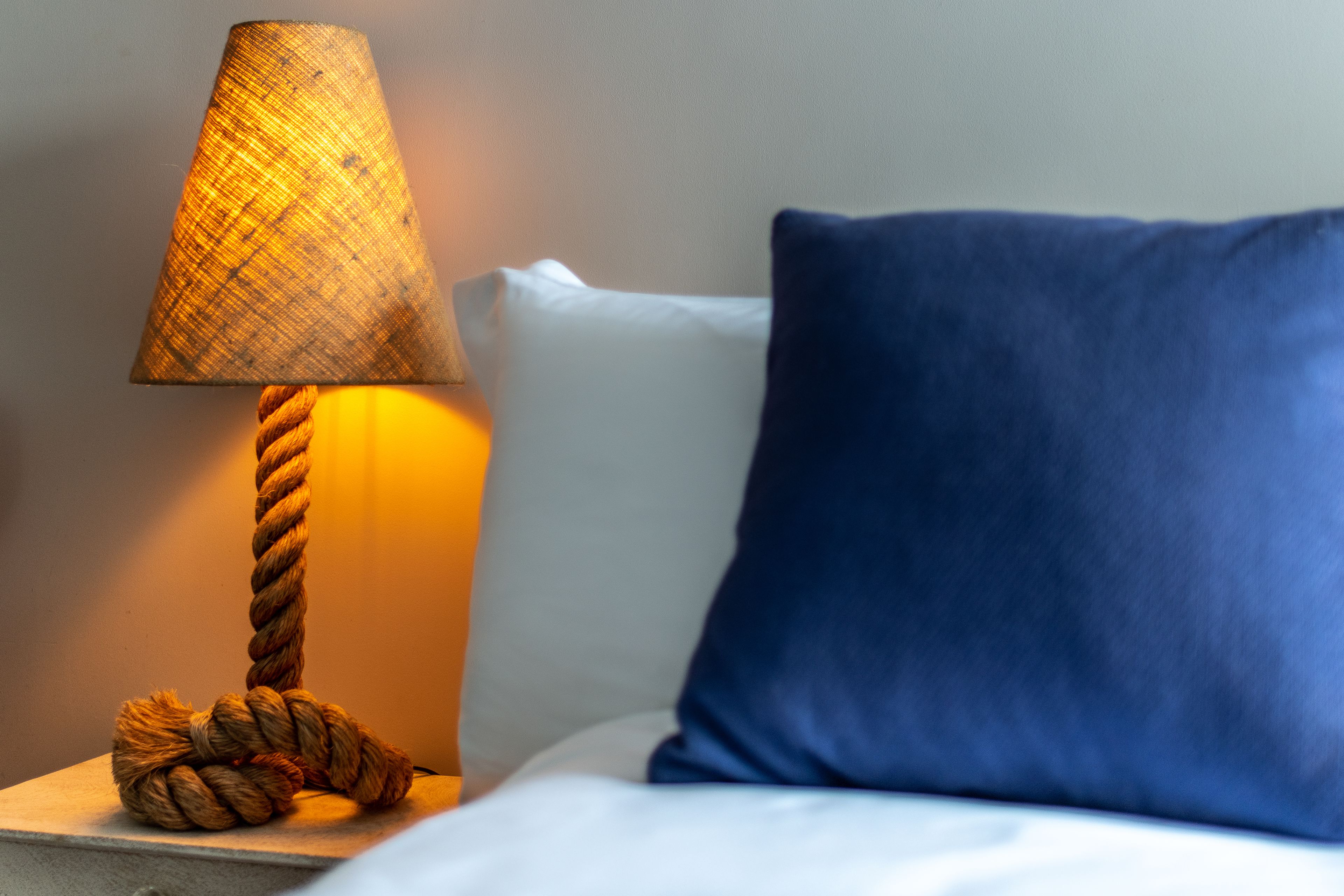 A bedside table with a lamp, a blue pillow, and a rope decoration.