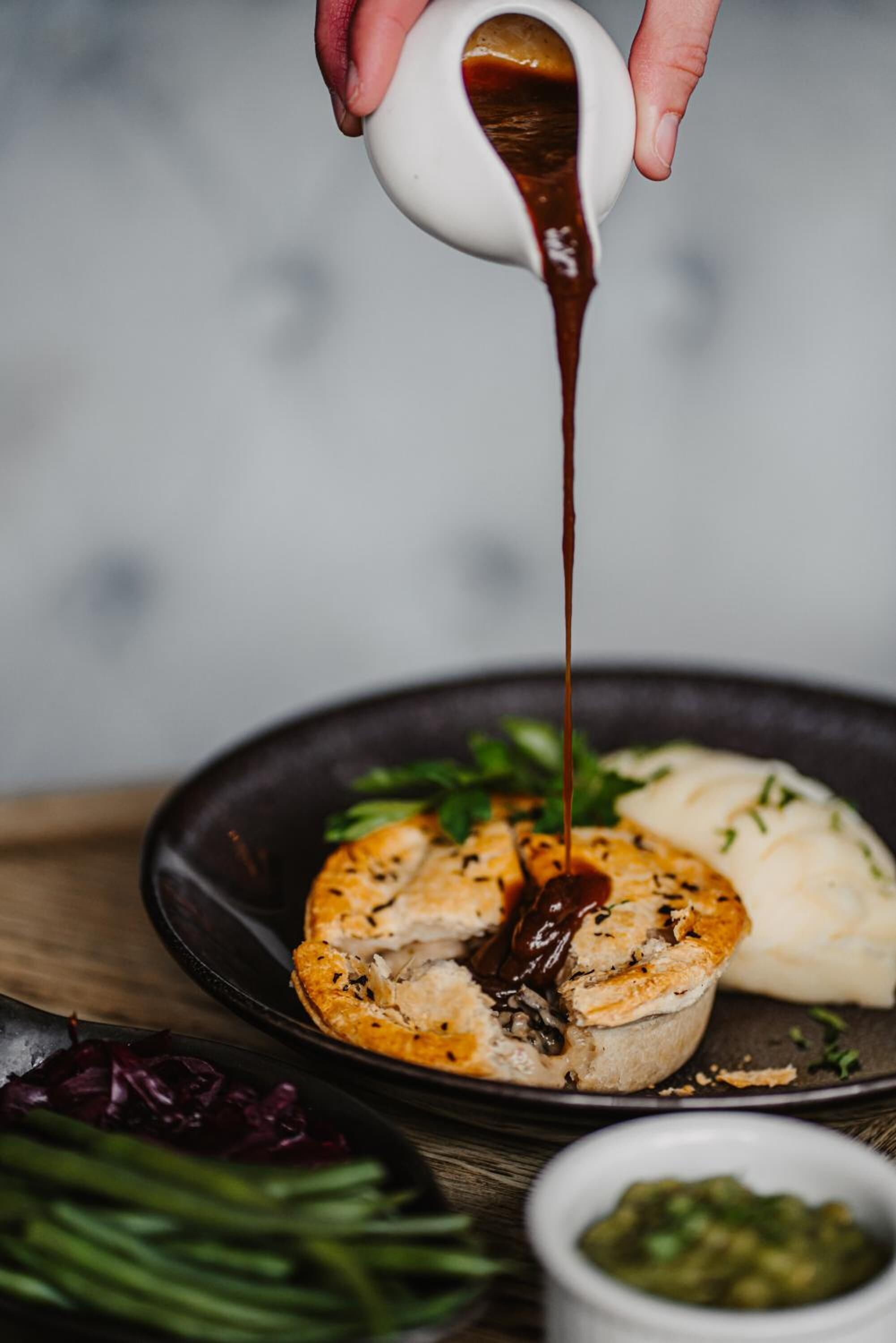 A hand pouring sauce over a dish with mashed potatoes and a piece of meat.
