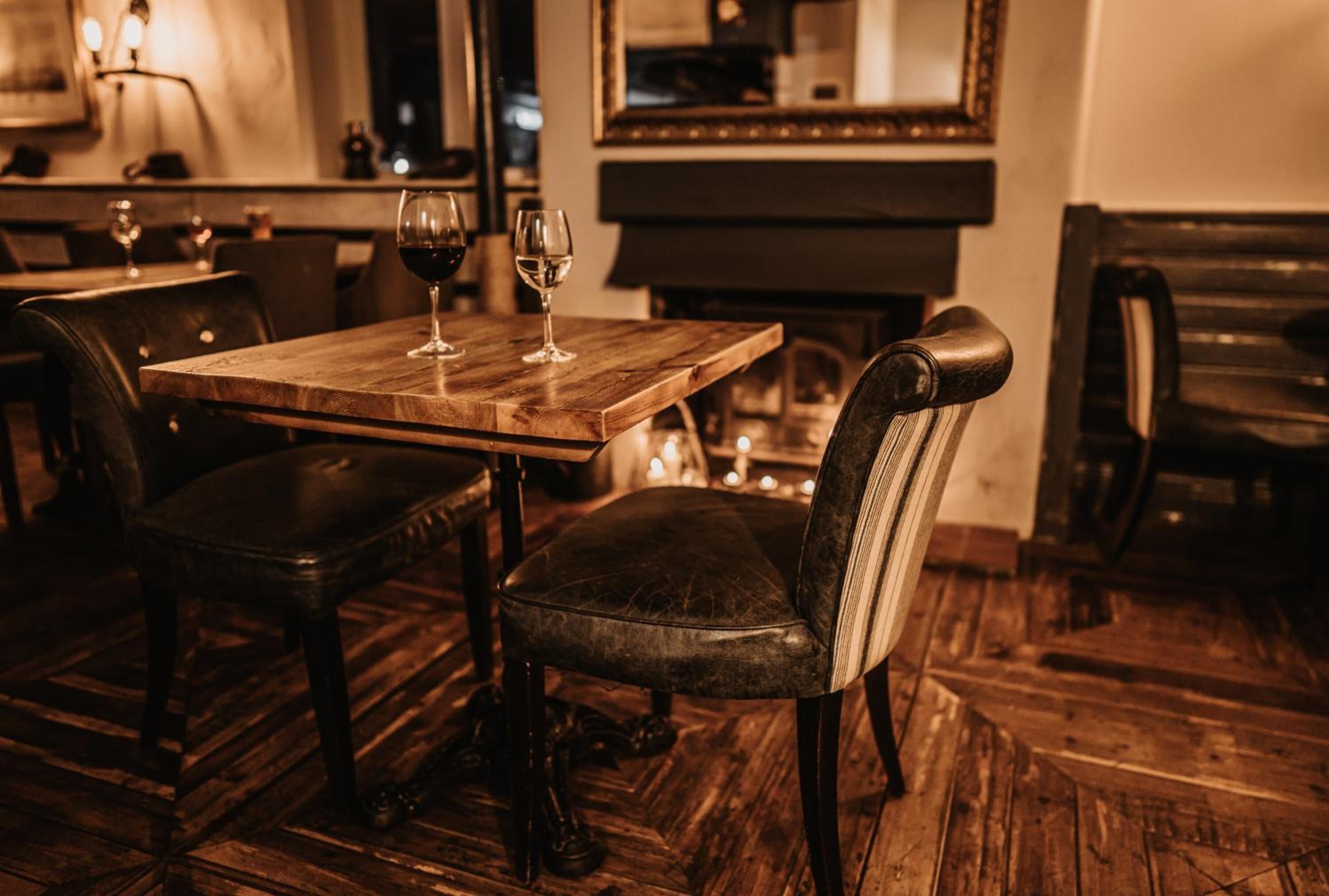 Ein Holztisch mit zwei Weingläsern und einem Stuhl mit Streifenmuster am Rücken, in einem schwach beleuchteten Raum mit Holzboden und einem Klavier im Hintergrund.