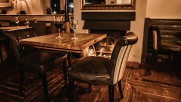 A wooden dining table with two wine glasses and a chair with a striped backrest in a dimly lit room with wooden flooring and a piano in the background.