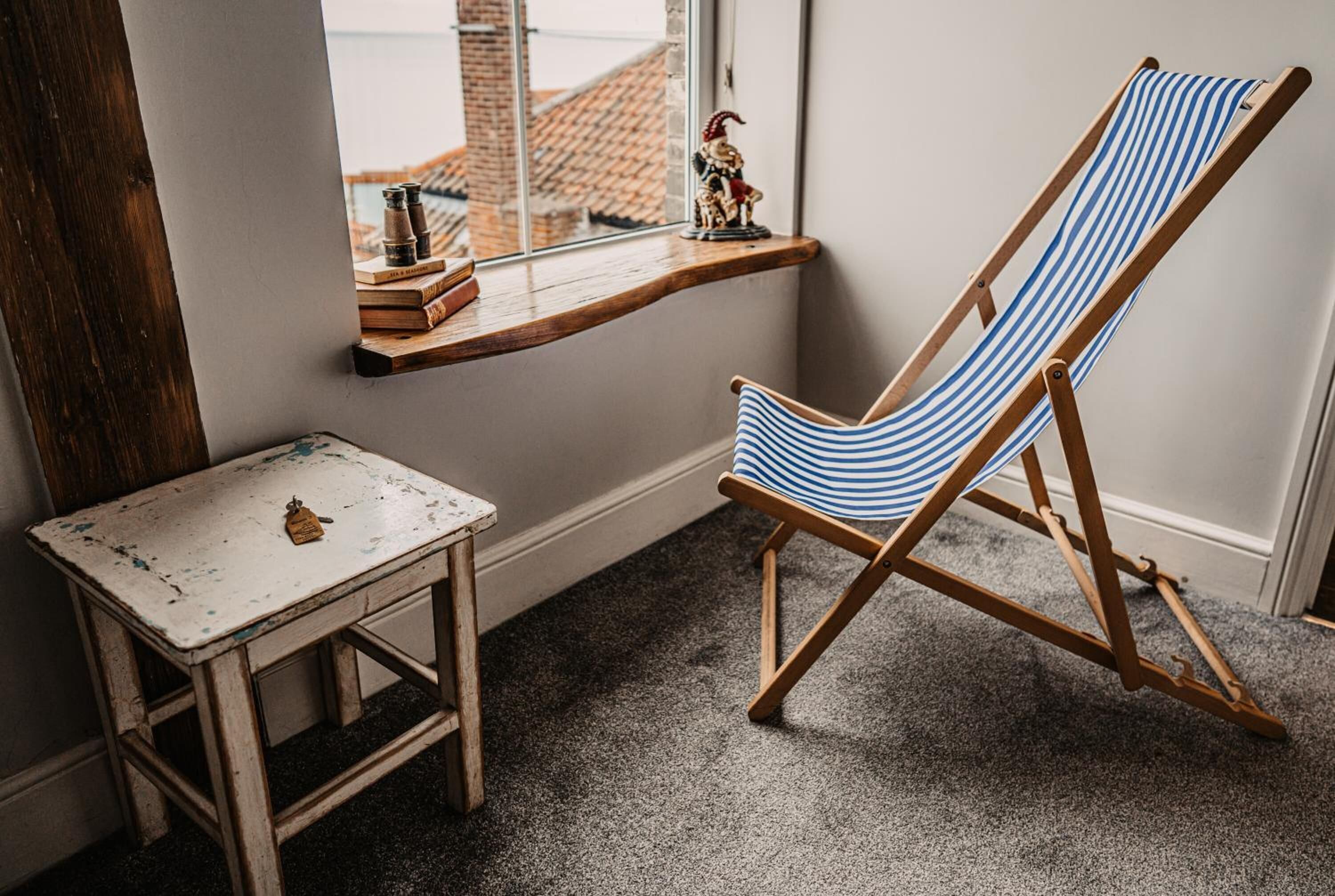 Una habitación con una silla de playa de madera y cojín a rayas, una pequeña mesa blanca con una campana de latón y una ventana con vistas a edificios.
