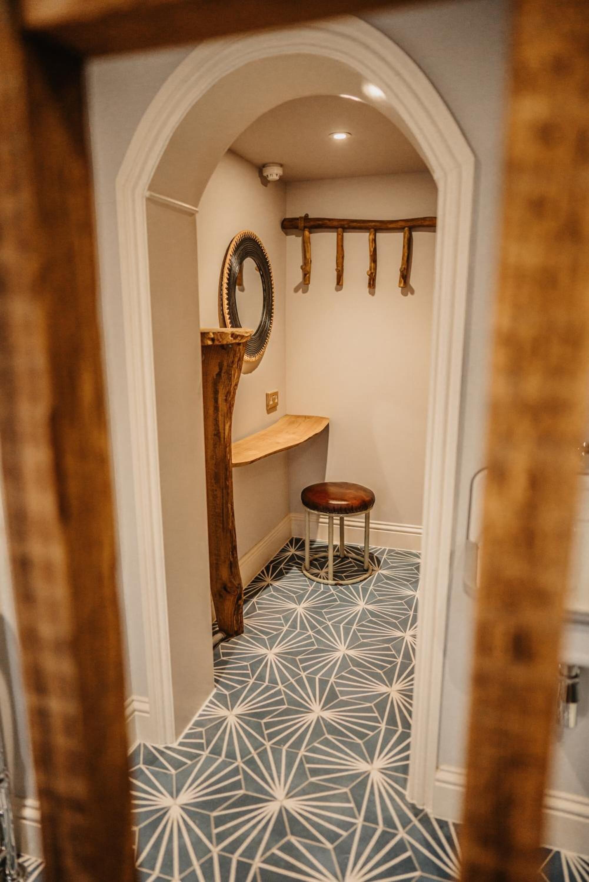 Un couloir étroit doté d’un sol à motifs géométriques, une colonne en bois, un miroir, un petit tabouret et un porte-manteau suspendu.