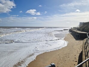 A beach with waves crashing onto the shore, a concrete walkway, and a building in the background.