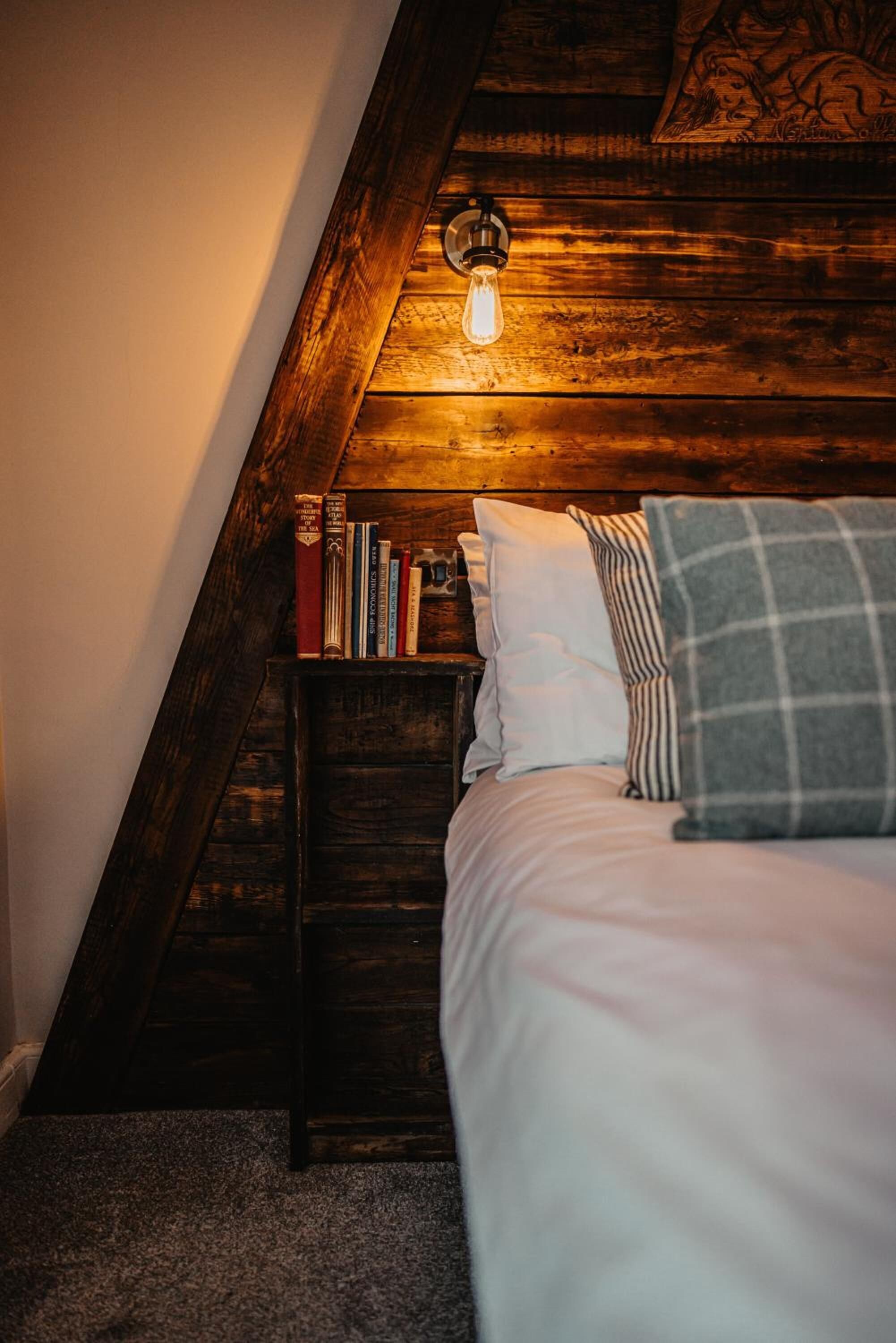Ein gemütliches Schlafzimmer mit schrägem Dach, einem hölzernen Kopfteil, einem Nachttisch mit Büchern, einer Lampe und einem Bett mit Kissen.