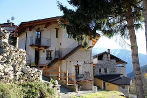 A two-story stone house with a wooden balcony, a stone staircase, and a mountain view.