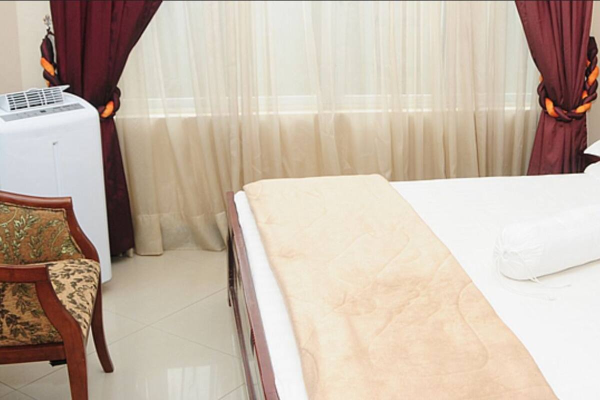 Una habitación de hotel con una cama, una silla, un ventanal con cortinas y un equipo de aire acondicionado.