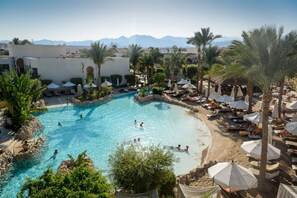 A resort pool with clear blue water, surrounded by palm trees and umbrellas, with mountains in the background. - Ghazala Gardens  (Sharm El Sheikh)