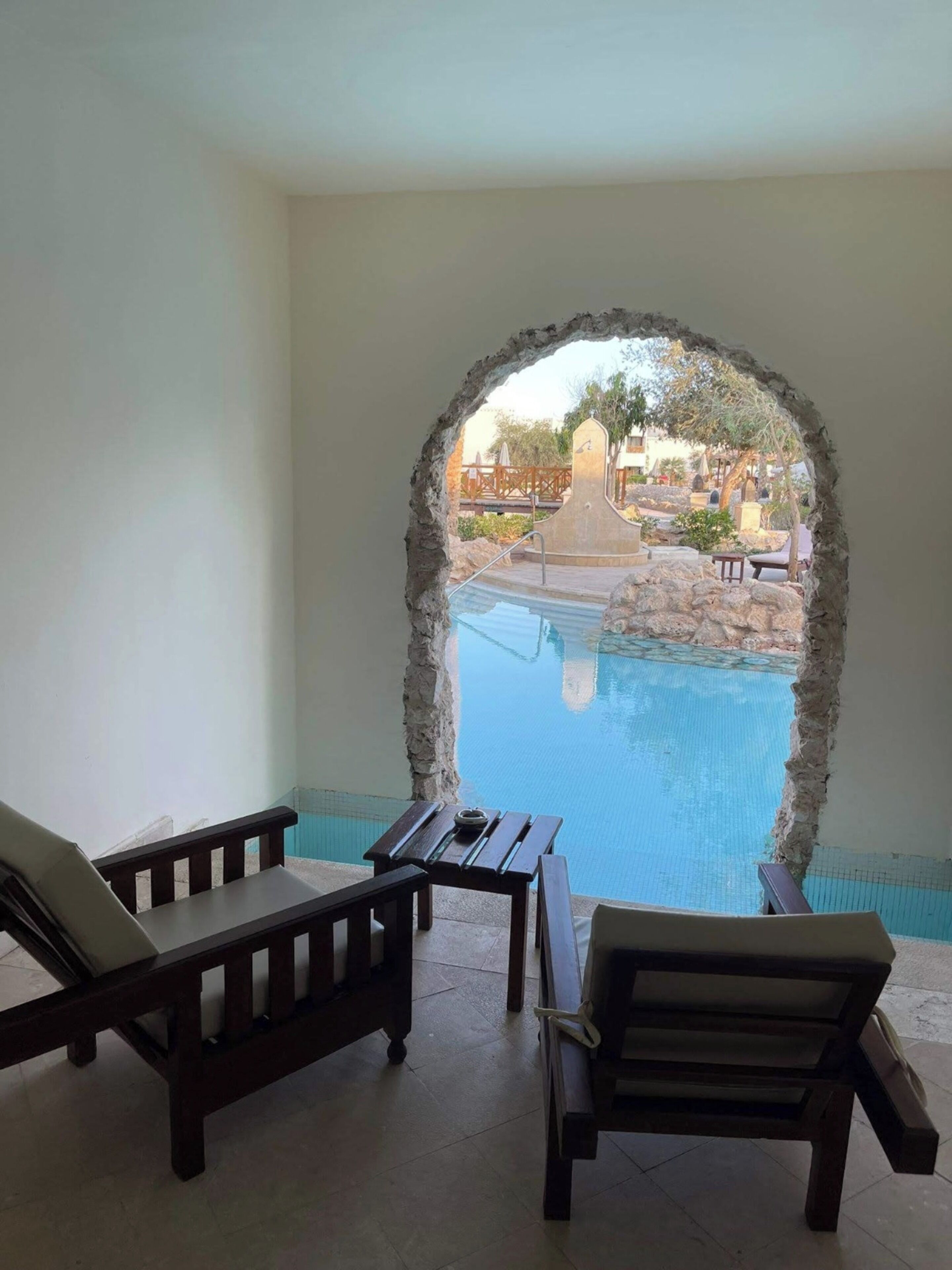 Un área de piscina con un arco de piedra, sillas de descanso y una pequeña mesa.