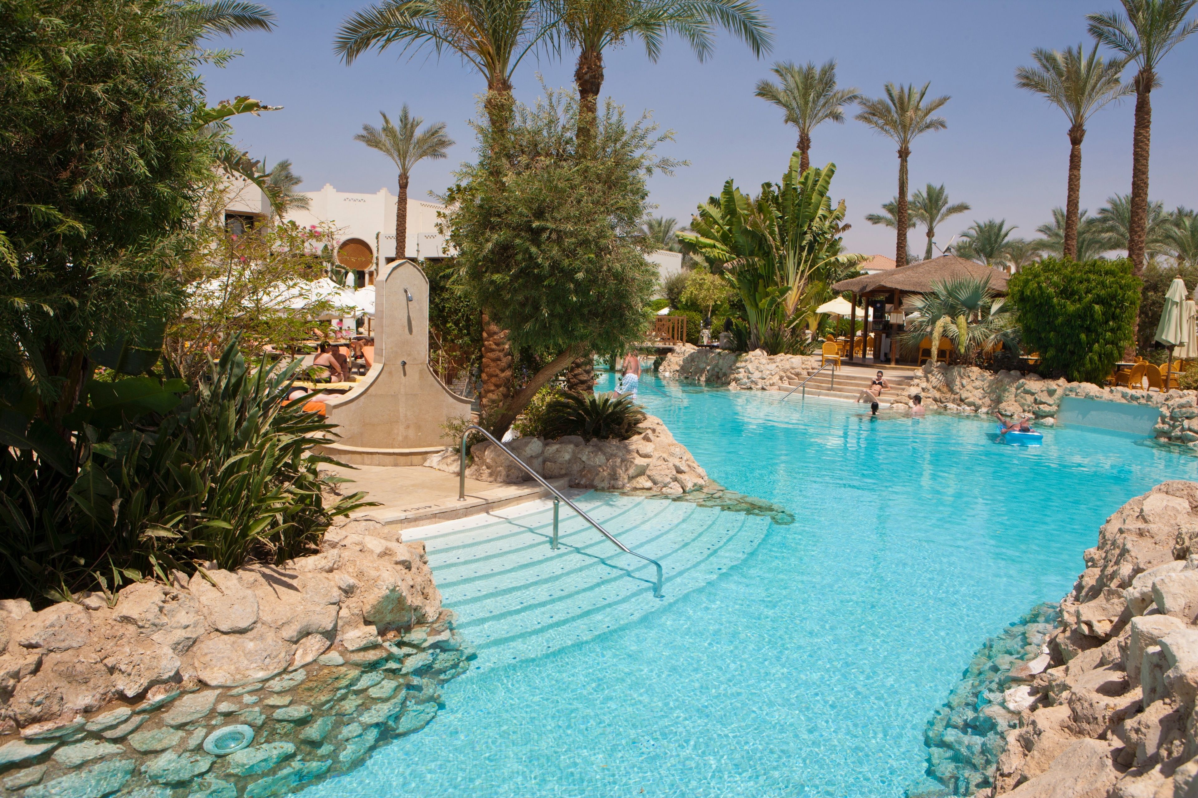 Zona de piscina con agua cristalina, rodeada de rocas y palmeras.