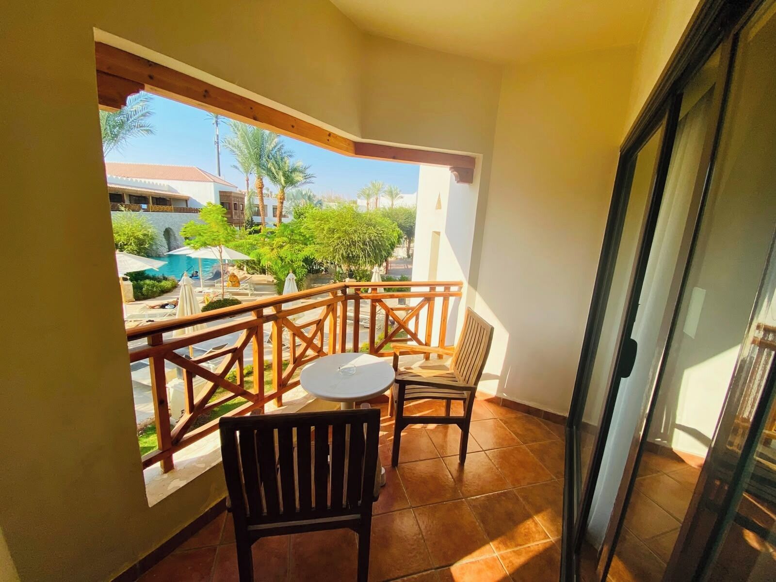 A balcony with a view of a pool and palm trees, a table, and two chairs.