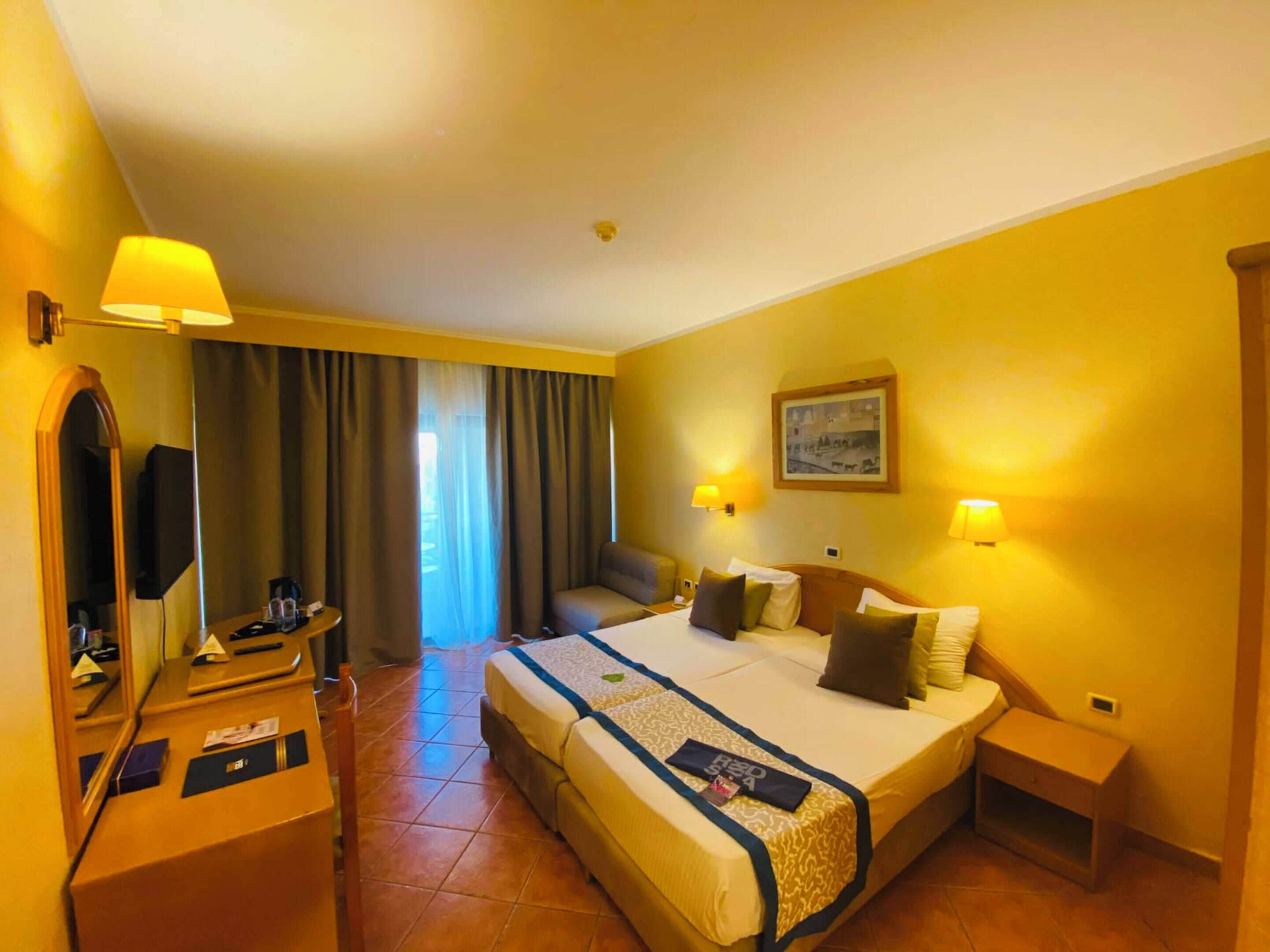 A hotel room with a bed, bedside lamps, a desk with a mirror, a television, and a window with curtains.