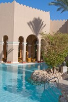 Un área de piscina con agua azul cristalina, una palmera y un edificio con arcos y columnas.
