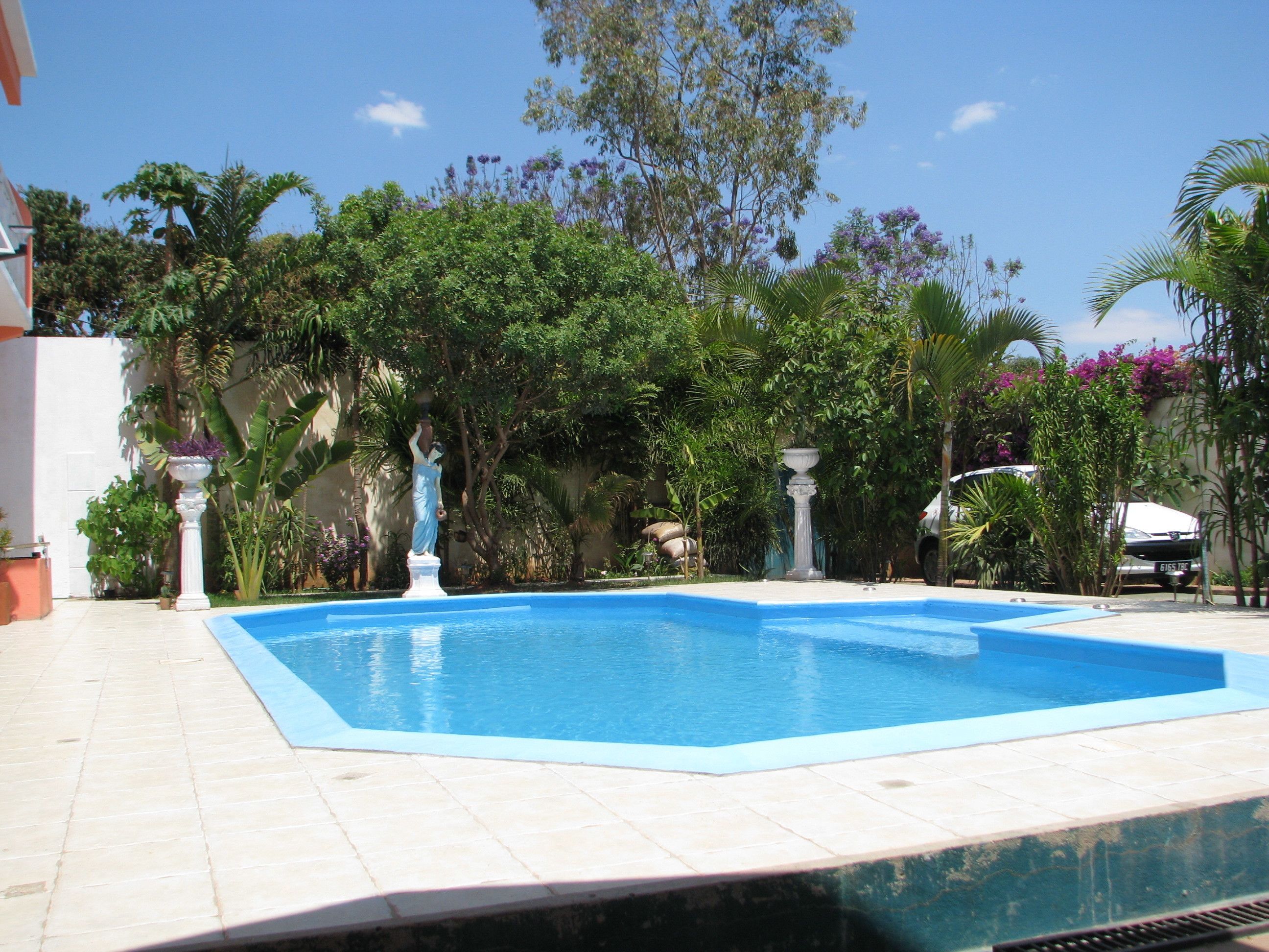 Una piscina con solárium de baldosas, rodeada de abundante vegetación y una estatua.