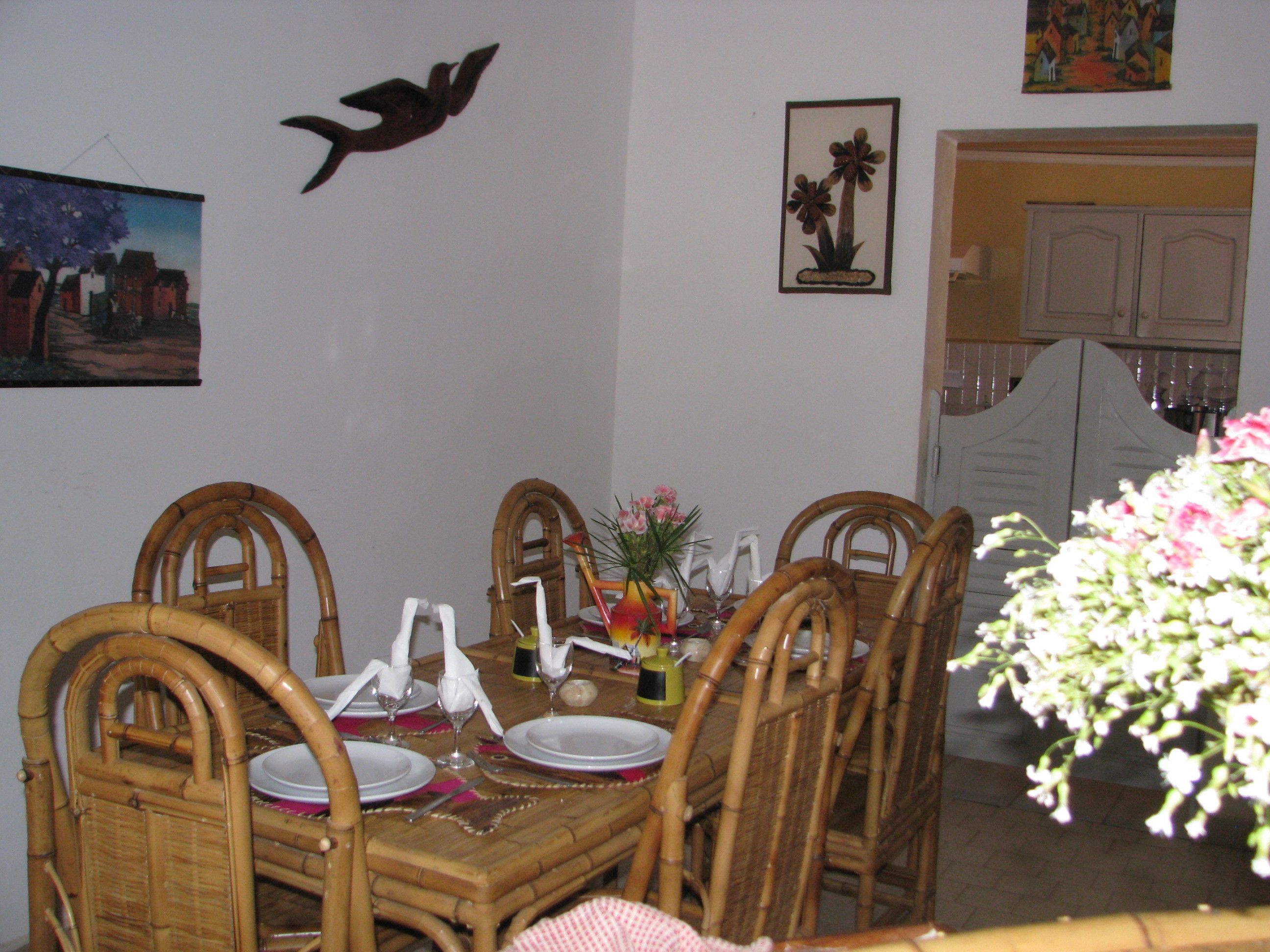 A dining area with a wooden table set for four, surrounded by wicker chairs. There are wall decorations including a bird and a framed picture.