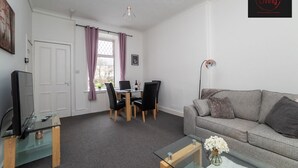 A modern living room with a grey sofa, a glass coffee table, a TV on a wooden stand, a dining area with a table and chairs, and a window with curtains. - One Bedroom Apartment by Klass Living Serviced Accommodation Coatbridge - Albion Apartment with Wifi and Parking (Coatbridge)