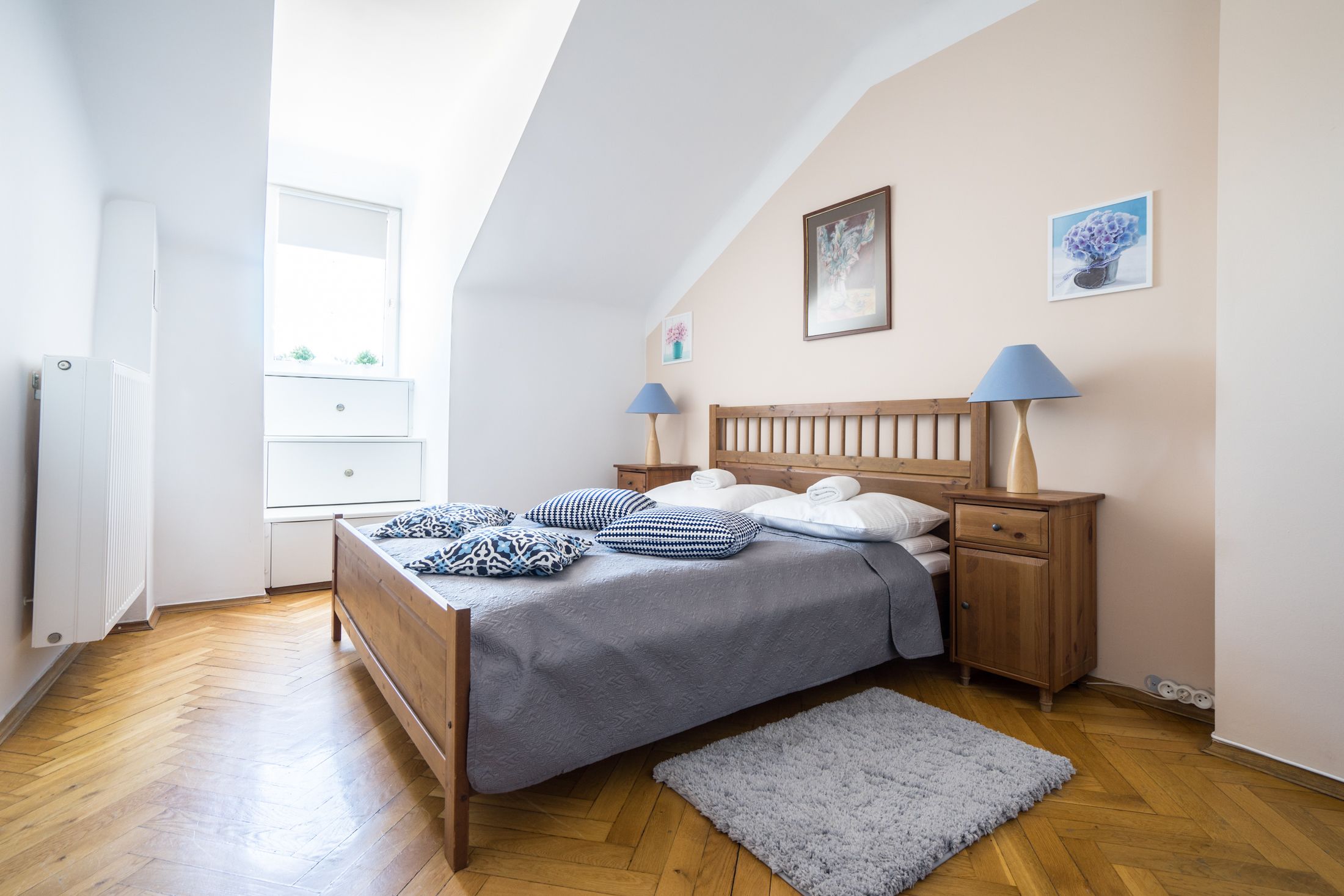 A bedroom with a wooden bed, a nightstand, two wall-mounted lamps, and a window with a view of greenery.
