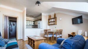 Ein modernes Wohnzimmer mit einer Couch, einem Esstisch mit Stühlen, einer Küche im Hintergrund und einem flachbildfernseher an der Wand.