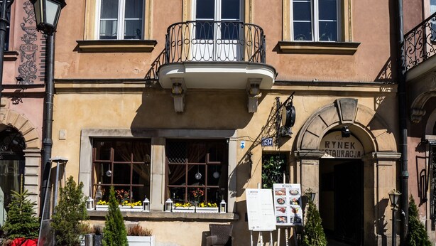 Ein Gebäude mit einem mittleren Eingang, einem Balkon mit schmiedeeisernem Geländer und einem Schild vor der Fassade.