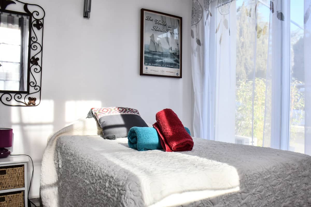 Un lit bien fait, recouvert d’une couvre-lit à motifs, avec deux oreillers et deux serviettes pliées. Un tableau encadré est accroché au mur, au-dessus du lit.