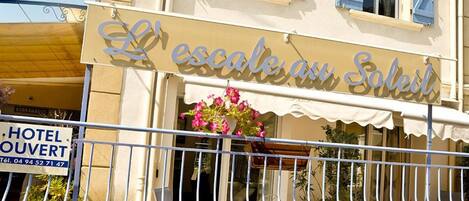 A hotel named "L'escalade au Soleil" with a balcony, outdoor seating, and a sign indicating it is a hotel.