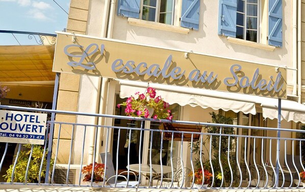 A hotel named "L'escalade au Soleil" with a balcony, outdoor seating, and a sign indicating it is a hotel.